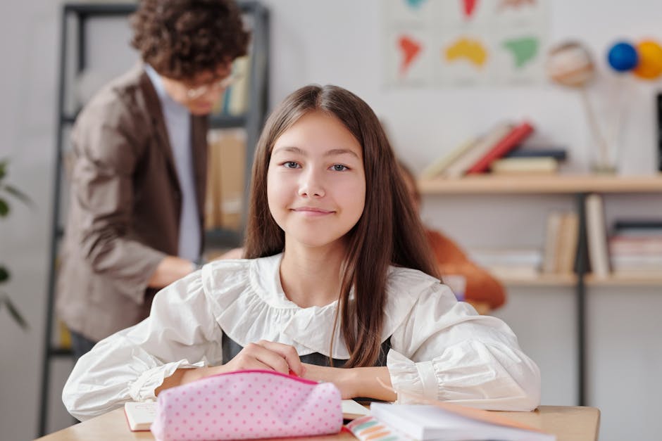 A young girl with long brown hair and a gentle smile sits at a desk in a well-lit indoor space, wearing a white blouse with puffy sleeves. In front of her are open notebooks and a pink, polka-dotted p